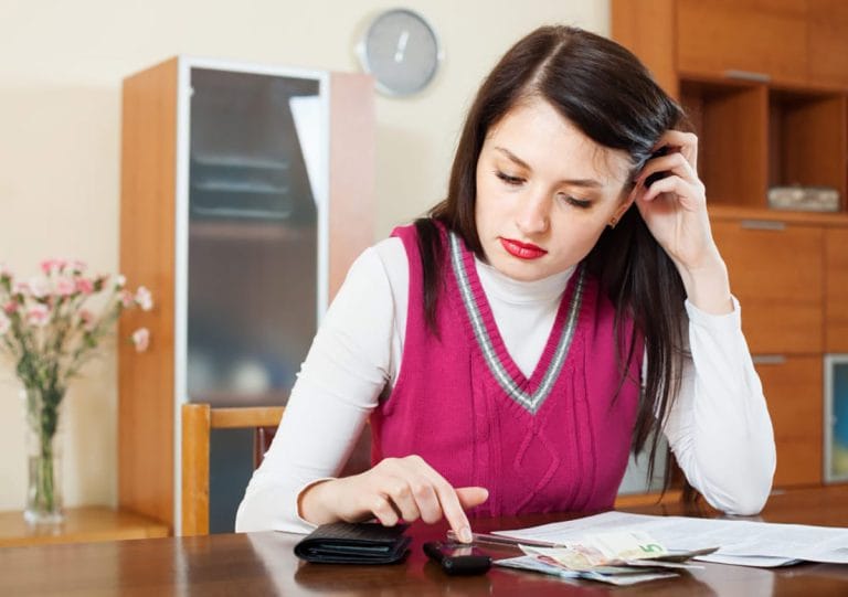 Woman sits at a wooden table, counting money with a calculator and receipts, looking stressed or worried about finances.”|